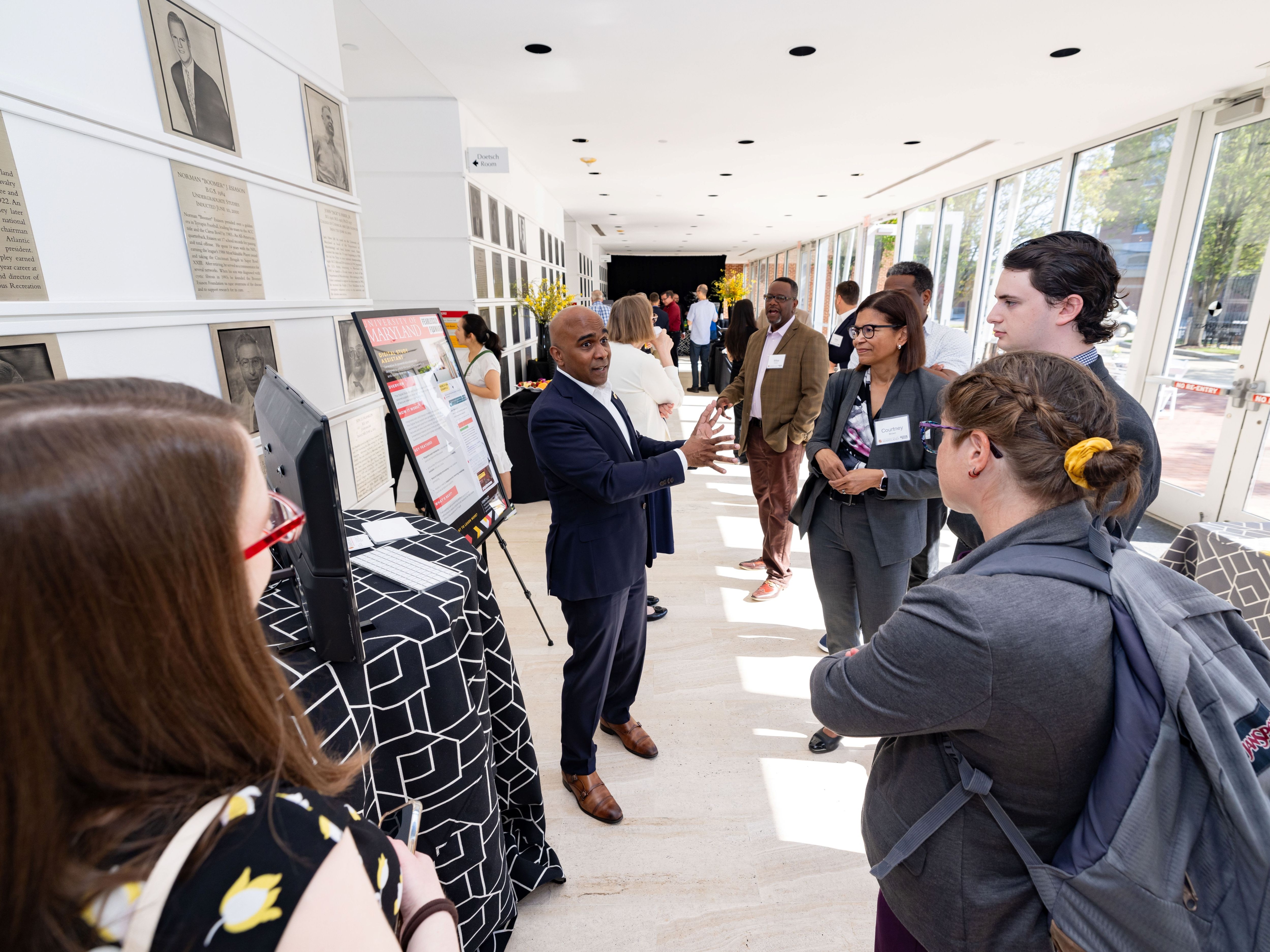 A man in a suit speaks to a small group of people gathered around a display in a bright hallway lined with portraits.